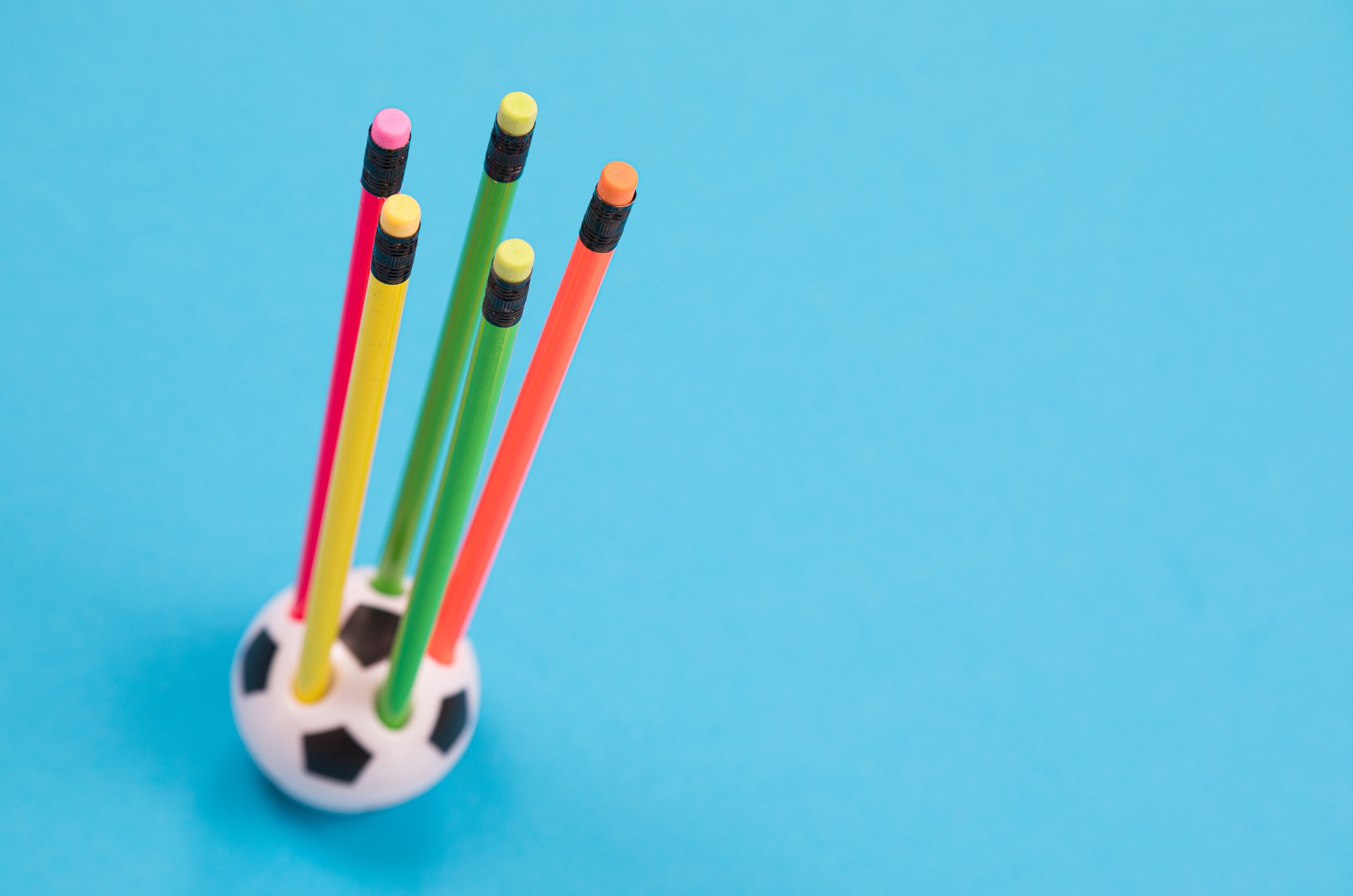 A round soccer ball shape pencil cup with five colorful pencils in it on azure surface isolated