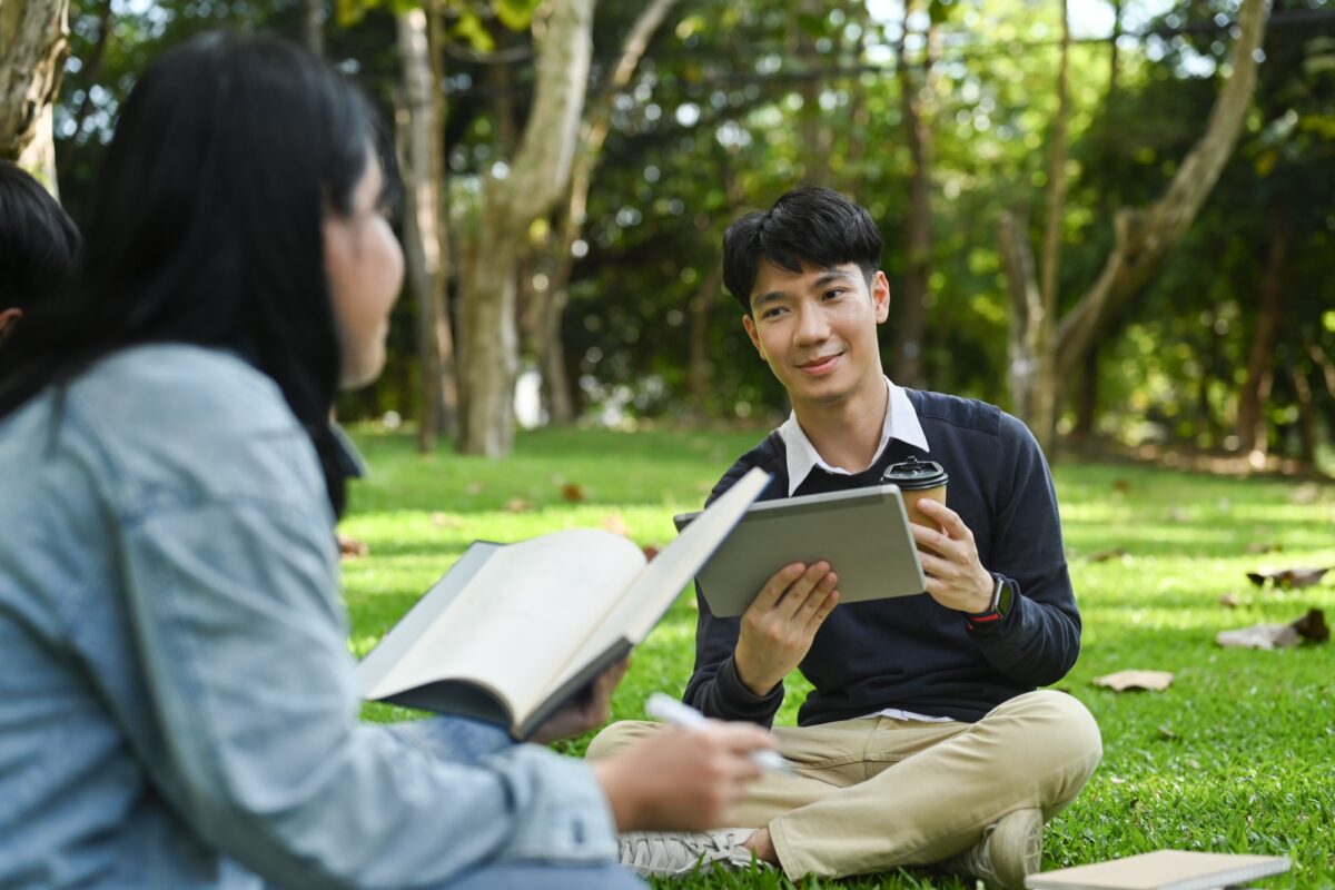 Smiling young asian male checking university schedule on digital tablet and talking with his friend.
