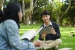 Smiling young asian male checking university schedule on digital tablet and talking with his friend.