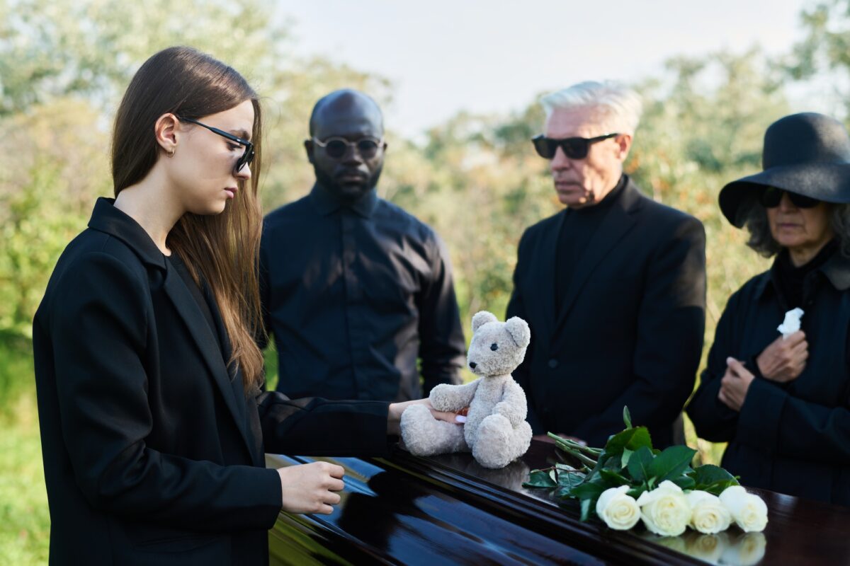 Young grieving woman putting grey teddybear on top of closed lid of coffin